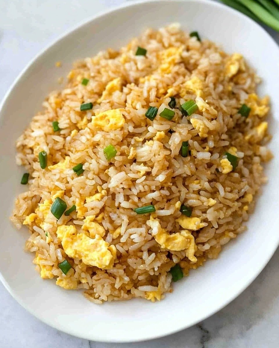 The image shows a close-up of a white ceramic spoon holding a scoop of fried rice with visible small scrambled egg pieces and finely chopped green onions mixed in. The rice is golden-brown with a slightly oily texture and small bits of egg spread throughout. Below the spoon, a large mound of the same fried rice fills a white plate, with the grains and egg pieces well mixed, resting on a white marbled surface. The colors are warm and inviting, mainly light brown from the fried rice, yellow from the eggs, and patches of green from the onions. Photo taken with an iphone --ar 4:5 --v 6.1
