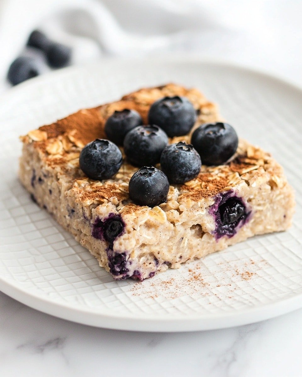 A thick, square-shaped baked oatmeal piece sits in a white ceramic dish with ornate edges, its top layer golden brown with a slightly rough texture. The oatmeal contains embedded dark blueberries, some of which have burst, leaving purple juices on the edges. A handful of whole blueberries are scattered on top, lightly dusted with a warm brown powder, most likely cinnamon. The dish is placed on a soft white marbled surface. Photo taken with an iphone --ar 4:5 --v 6.1