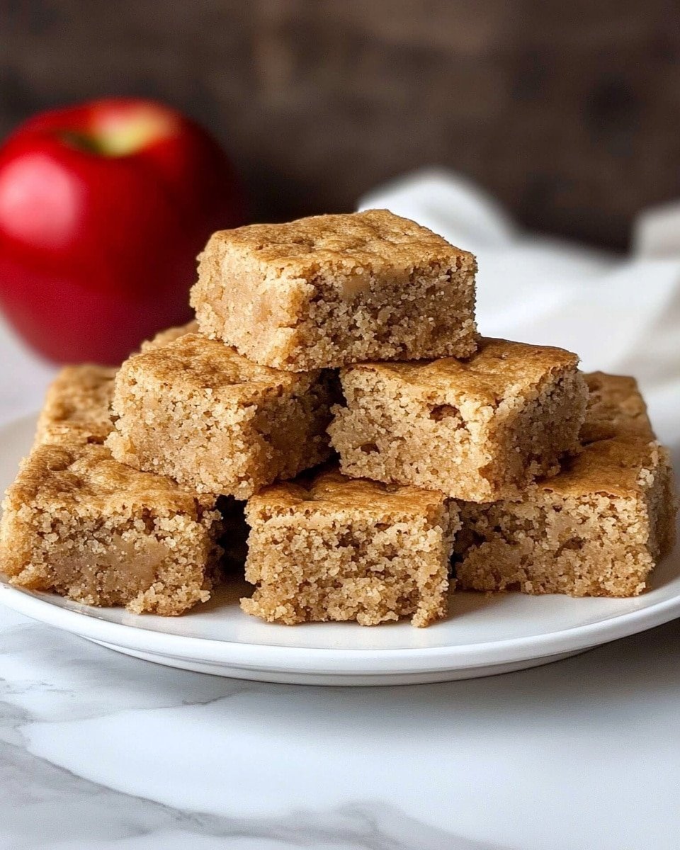 A white plate holds a stack of nine brown squares of soft cake arranged in three layers: the bottom layer has five pieces lying flat, the middle layer has three pieces stacked on top, and the top layer has one piece resting in the center. The cake has a crumbly texture with small bits inside, showing a mix of light and medium brown colors. A whole red apple is blurred in the background to the left, and the surface beneath the plate is a white marbled texture. Photo taken with an iphone --ar 4:5 --v 6.1