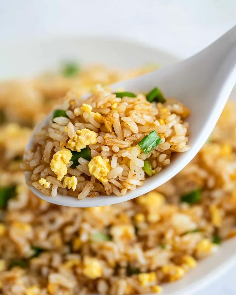 A close-up of a white ceramic spoon holding a serving of fried rice with visible grains of light brown rice mixed with small pieces of yellow scrambled egg and green onion bits. Below the spoon, a white plate is filled with more of the same fried rice, showing a mix of fluffy rice grains, yellow scrambled egg pieces, and scattered green onion bits. The background is a white marbled texture, making the colors of the fried rice stand out clearly. photo taken with an iphone --ar 4:5 --v 6.1