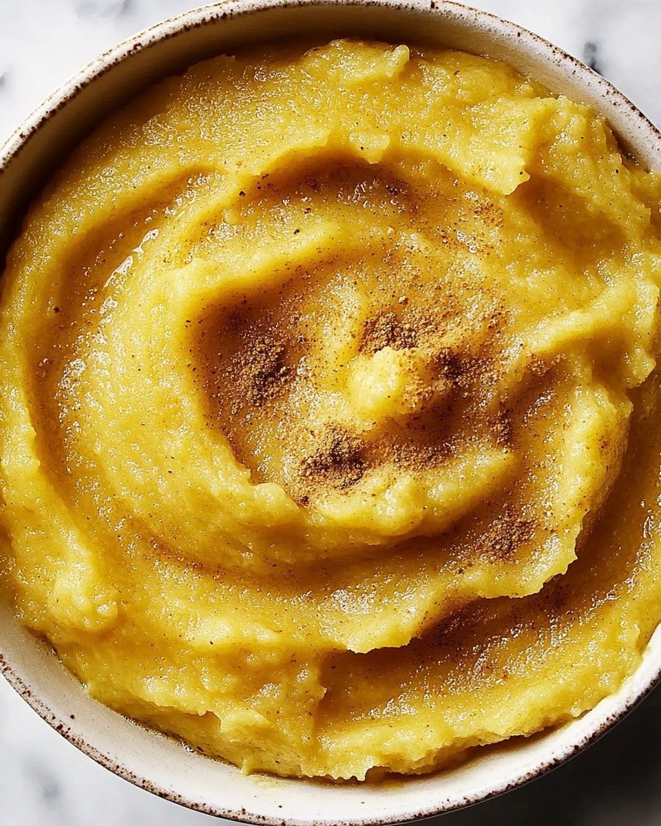 A close-up view of a bowl filled with smooth, thick mashed yellow food with a slightly grainy texture. The mash swirls around the bowl with some peaks and valleys showing its creamy consistency. Light brown spices are sprinkled evenly on top, adding contrast to the yellow base. The bowl is white and set on a white marbled surface, showing texture and slight shadows. The scene is well lit, highlighting the softness and rich texture of the dish. photo taken with an iphone --ar 4:5 --v 6.1